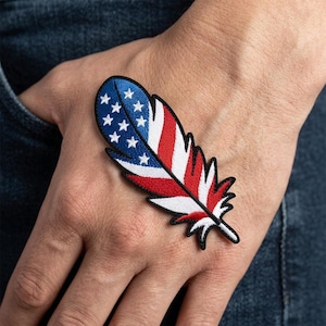 May include: An embroidered patch in the shape of a feather featuring the American flag design. The top section is blue with white stars, while the lower portion displays alternating red and white stripes, all outlined with a black stitched border.