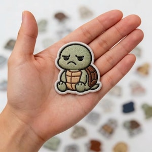 May include: An embroidered patch featuring a cartoon turtle with a grumpy facial expression. The turtle has a light green head and limbs, a tan-colored belly, and a brown shell, all outlined with a thick white border against a fabric backing.
