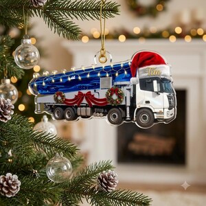 May include: A flat acrylic Christmas tree ornament shaped like a white concrete pump truck. The truck features a red Santa hat on the cab with the name Tim written in gold, a blue boom arm, red bows, and green wreaths. A string of lights is printed on top.