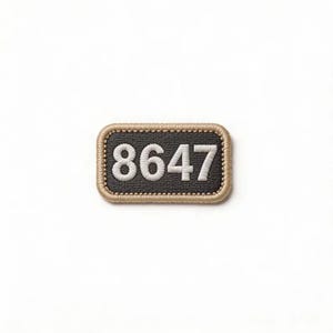 May include: A rectangular embroidered patch featuring the white numbers 8647 centered on a dark charcoal gray background. The patch is finished with a thick, tan-colored stitched border that frames the rectangular shape with rounded corners.