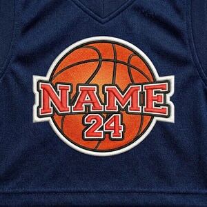May include: Navy blue basketball jersey with a large orange basketball graphic. The graphic includes the word "NAME" above the number "24" in red, outlined in white. The jersey has a V-neck.