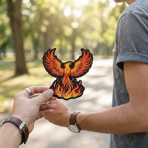 May include: Embroidered patch featuring a phoenix design in shades of orange, red, and yellow, with black outlining. The phoenix is depicted with outstretched wings and flames at its base. This patch is suitable for jackets, bags, or other fabric items.