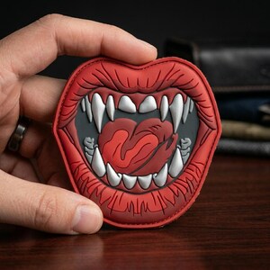 May include: A red patch featuring a stylized open mouth with sharp white fangs and a red tongue. The mouth is outlined in black and red, with a gray interior. The patch is held in a hand.