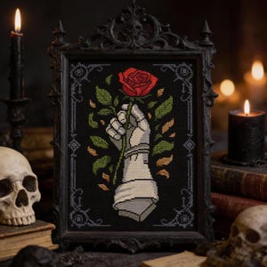 May include: Cross-stitch art in a black frame. The artwork depicts a white, armored hand holding a red rose against a black background. Green leaves and small orange accents surround the hand. The frame has a decorative, gothic design.