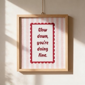May include: A framed cross-stitch artwork with the words "Slow down, you're doing fine." The text is in red on a white background, framed by a red scalloped border. The background has pink and white vertical stripes, and the frame is light wood.
