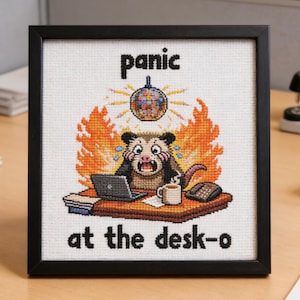 Op de afbeelding: Een ingelijste kruissteekkunst in een zwart frame. Het kunstwerk toont een gestreste opossum aan een bureau, omringd door vlammen en een discobal. Tekst luidt "panic at the desk-o". Het kleurenpalet omvat oranje, zwart, wit en bruin.