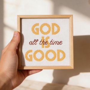 Puede incluir: Una obra de arte en punto de cruz enmarcada con las palabras "GOD is all the time GOOD" en hilo amarillo y granate. La obra de arte está sobre un fondo blanco y enmarcada en un marco de madera marrón claro. La pieza está sujeta por una mano.
