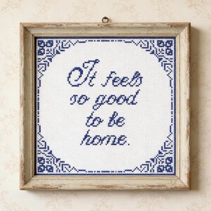 Peut inclure: Une œuvre d'art encadrée en point de croix avec le texte "It feels so good to be home." Le texte est en fil bleu foncé sur fond blanc, entouré d'une bordure bleue à motifs. Le cadre est en bois clair vieilli.