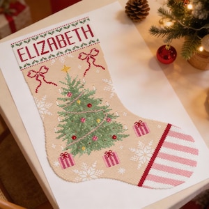 May include: A beige Christmas stocking with the name "ELIZABETH" embroidered in red. The stocking features a green Christmas tree, pink gift boxes, and snowflakes. A red and white striped pattern accents the toe.