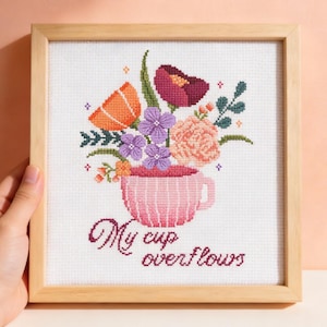 May include: Cross-stitch artwork in a wooden frame. The design shows a pink teacup overflowing with colourful flowers, including orange, purple, and peach blooms. The text "My cup overflows" is stitched below the cup in a cursive font.