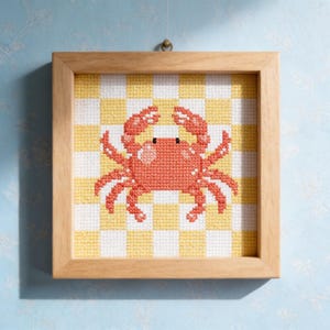 Puede incluir: Una obra de arte en punto de cruz enmarcada con un cangrejo de color coral sobre un fondo a cuadros amarillo y blanco. El cangrejo tiene ojos negros y está enmarcado en un marco de madera natural, ideal para la decoración costera.