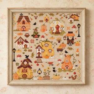 May include: A framed cross-stitch sampler on beige fabric featuring a collection of autumn-themed motifs including pumpkin-shaped houses, small carts, mushrooms, black cats, a squirrel, and street lamps, all rendered in shades of orange, brown, and green.