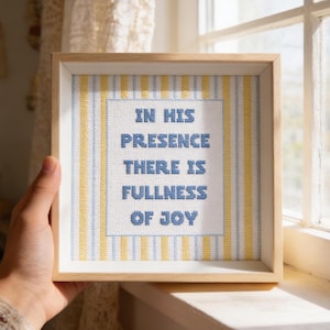May include: A framed cross-stitch artwork with the text "IN HIS PRESENCE THERE IS FULLNESS OF JOY" in blue thread. The background features vertical stripes in yellow and light blue, encased in a light wood frame.