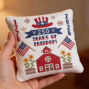 May include: A small, square, white cross-stitch pillow with patriotic imagery. It features a red barn, American flags, and the text "250 Years of Freedom" with the years 1776 and 2026. The design includes a red, white, and blue Uncle Sam hat.