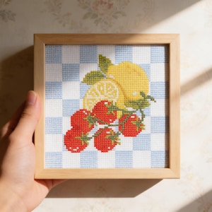 Könnte beinhalten: Ein gerahmtes Kreuzstich-Kunstwerk mit einer Zitrone und Tomaten. Die Zitrone ist gelb mit grünen Blättern, und die Tomaten sind rot. Der Hintergrund ist ein blau-weißes Karomuster, alles in einem hellbraunen Holzrahmen.