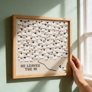 Könnte beinhalten: Gerahmtes Kreuzstich-Kunstwerk mit einer Herde weißer Schafe mit schwarzen Gesichtern und Beinen auf beigem Hintergrund. Der Text "HE LEAVES THE 99" ist darunter gestickt. Der Rahmen ist aus hellem Holz.
