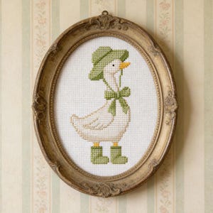 Pode incluir: Obra de arte em ponto cruz de um ganso usando chapéu, laço e botas verdes, em uma moldura oval dourada e ornamentada. O ganso tem um bico amarelo e está em um fundo branco. A moldura está pendurada em uma parede listrada.