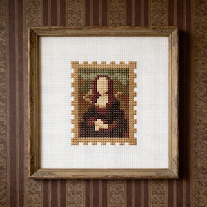 Könnte beinhalten: Kreuzstich-Kunstwerk der Mona Lisa, gerahmt in einem quadratischen, hellbraunen Holzrahmen. Das Kunstwerk ist eine detaillierte, pixelige Darstellung des berühmten Gemäldes, mit einem neutralen Hintergrund. Der Rahmen ist auf einer braun-beigen gestreiften Tapete montiert.