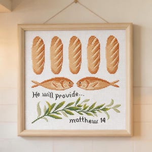 Op de afbeelding: Kruissteekkunstwerk in een licht houten frame. Het ontwerp toont zes broden, twee vissen en een takje bladeren. De tekst luidt "He will provide... matthew 14". Het kunstwerk staat op een witte achtergrond.