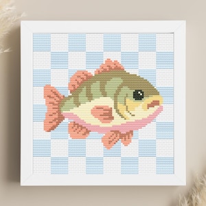 May include: A framed cross-stitch of a fish, with a light green body, pink and peach details, and a black eye. The background is a blue and white chequered pattern. The frame is white, creating a clean, simple aesthetic.