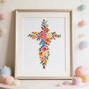 Peut inclure: Une œuvre d'art au point de croix encadrée, représentant une croix florale. La croix est ornée de fleurs roses, oranges, jaunes et bleues sur fond blanc. Une décoration pour la maison.