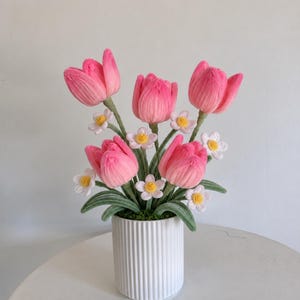 Pode incluir: Um buquê de tulipas cor-de-rosa de crochê e pequenas flores brancas com centros amarelos, com caules e folhas verdes, disposto num vaso cilíndrico de cerâmica branca com detalhes estriados verticais na superfície exterior.