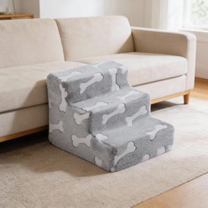 Puede incluir: Una escalera para mascotas de tres escalones en tejido gris suave con un patrón de huesos blancos. Las escaleras están diseñadas para ayudar a perros o gatos pequeños a alcanzar superficies más altas como un sofá. Las escaleras están colocadas sobre una alfombra de color neutro.
