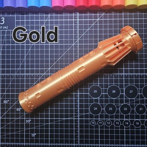 Pu&ograve; includere: Un oggetto cilindrico color oro con dettagli intricati, possibilmente un oggetto di scena o decorativo. La parola "Gold" &egrave; stampata in bianco in grassetto. L'oggetto &egrave; su un tappetino da taglio nero con segni di misurazione e pennarelli colorati sullo sfondo.