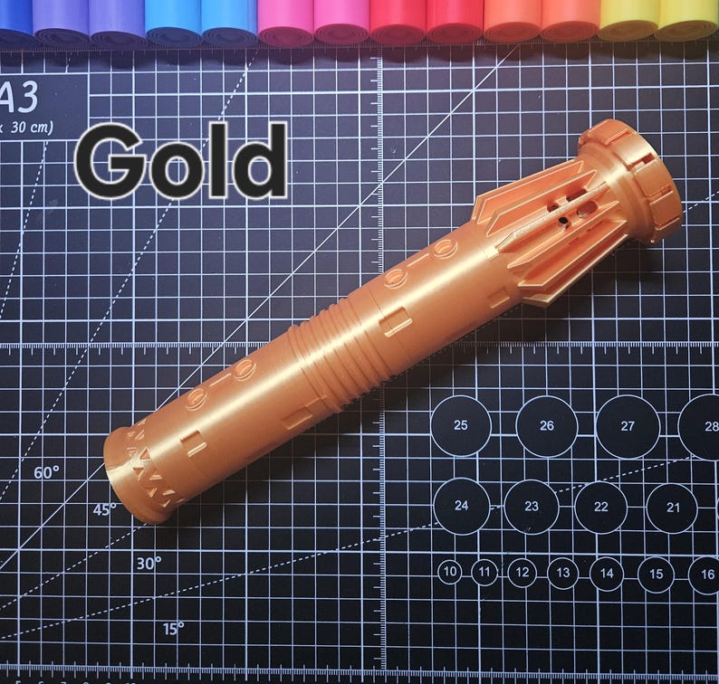 Pu&ograve; includere: Un oggetto cilindrico color oro con dettagli intricati, possibilmente un oggetto di scena o decorativo. La parola "Gold" &egrave; stampata in bianco in grassetto. L'oggetto &egrave; su un tappetino da taglio nero con segni di misurazione e pennarelli colorati sullo sfondo.