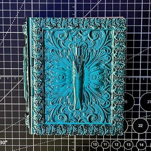 May include: A teal-colored, ornate box with a detailed floral and geometric design. The box features a central motif resembling a stylized arrow or feather, surrounded by symmetrical patterns. The box is likely made of a hard material and has a hinged design.