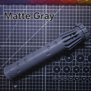 Pu&ograve; includere: Un'elsa di spada laser grigio opaco, con un design cilindrico dettagliato e accenti circolari e rettangolari. Il testo "Matte Gray" &egrave; visualizzato sopra. Lo sfondo &egrave; un tappetino da taglio nero con segni di misurazione.