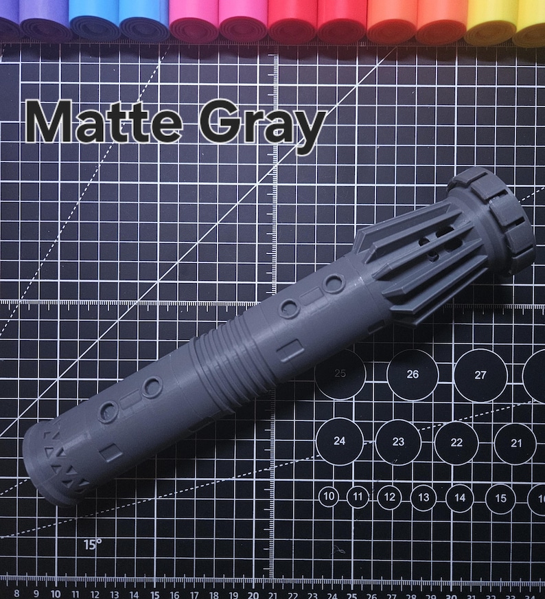 Pu&ograve; includere: Un'elsa di spada laser grigio opaco, con un design cilindrico dettagliato e accenti circolari e rettangolari. Il testo "Matte Gray" &egrave; visualizzato sopra. Lo sfondo &egrave; un tappetino da taglio nero con segni di misurazione.