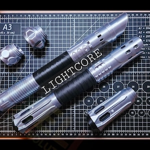May include: Two silver and black lightsaber hilts with metallic finishes and textured grips. One hilt is fully assembled, while the other is shown with two separate silver pommel or emitter components placed nearby. The word LIGHTCORE is visible.