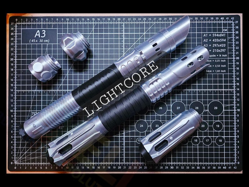 May include: Two silver and black lightsaber hilts with metallic finishes and textured grips. One hilt is fully assembled, while the other is shown with two separate silver pommel or emitter components placed nearby. The word LIGHTCORE is visible.