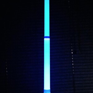 May include: A vertical, cylindrical light wand emitting a bright blue glow, featuring segmented sections of varying intensity. The base is dark, and the word LIGHTCORE is printed in white, capitalised, serif font at the bottom of the image.
