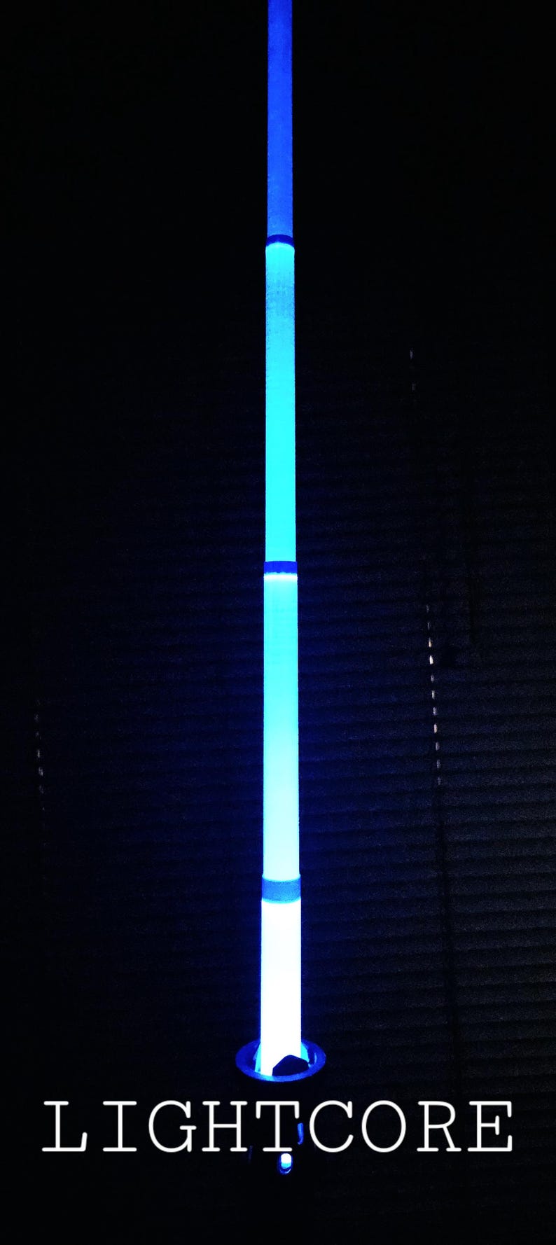 May include: A vertical, cylindrical light wand emitting a bright blue glow, featuring segmented sections of varying intensity. The base is dark, and the word LIGHTCORE is printed in white, capitalised, serif font at the bottom of the image.