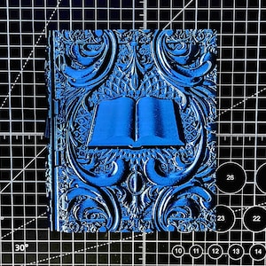 May include: A blue, embossed book-shaped object with an open book design in the center. The cover features ornate, swirling patterns. The object is on a black cutting mat with white grid lines and numbers.