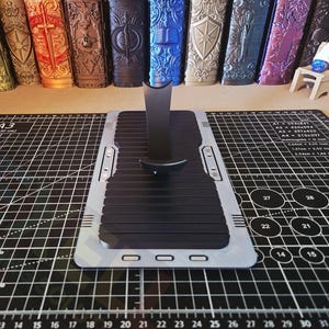 Puede incluir: Un soporte para tel&eacute;fono negro y plateado con una superficie acanalada, colocado sobre una esterilla de corte negra con l&iacute;neas de cuadr&iacute;cula y medidas. Varios objetos decorativos est&aacute;n en el fondo.