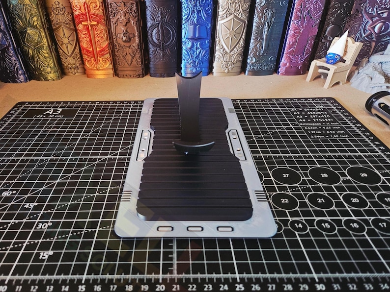 Puede incluir: Un soporte para tel&eacute;fono negro y plateado con una superficie acanalada, colocado sobre una esterilla de corte negra con l&iacute;neas de cuadr&iacute;cula y medidas. Varios objetos decorativos est&aacute;n en el fondo.