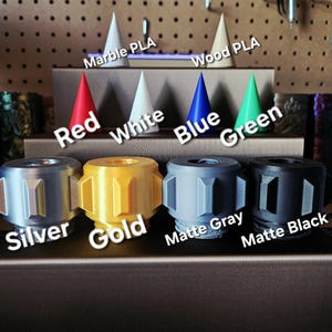 Puede incluir: Una exhibici&oacute;n de objetos impresos en 3D en varios colores y materiales. La imagen muestra varios objetos cil&iacute;ndricos en plata, oro, gris mate y negro mate, junto con objetos en forma de cono en rojo, blanco, azul y verde. Las etiquetas de texto identifican los colores y materiales.
