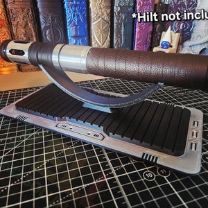 Puede incluir: Un soporte de exhibici&oacute;n para empu&ntilde;adura de sable de luz con una base negra y plateada, que sostiene una empu&ntilde;adura de sable de luz marr&oacute;n y plateada. El soporte tiene un soporte curvo y una base rectangular con barras negras. El texto "*Hilt not included*" es visible.