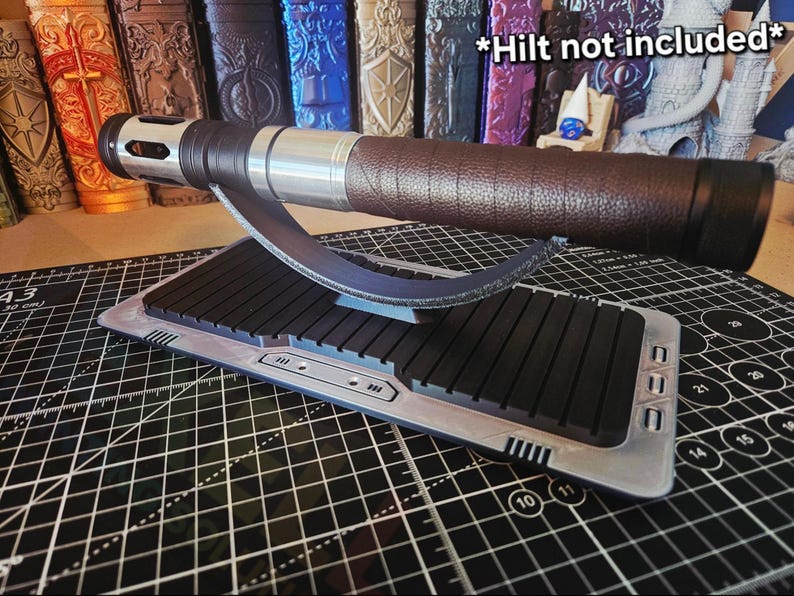 Puede incluir: Un soporte de exhibici&oacute;n para empu&ntilde;adura de sable de luz con una base negra y plateada, que sostiene una empu&ntilde;adura de sable de luz marr&oacute;n y plateada. El soporte tiene un soporte curvo y una base rectangular con barras negras. El texto "*Hilt not included*" es visible.