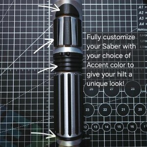 May include: A silver and black cylindrical lightsaber hilt featuring multiple textured grip sections and accent rings. The text on the image reads: Fully customize your Saber with your choice of Accent color to give your hilt a unique look!