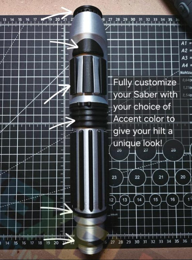 May include: A silver and black cylindrical lightsaber hilt featuring multiple textured grip sections and accent rings. The text on the image reads: Fully customize your Saber with your choice of Accent color to give your hilt a unique look!