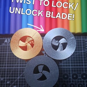 Pu&ograve; includere: Tre oggetti circolari metallici con un design a forma di ventaglio in oro, argento e grigio scuro. Il testo "TWIST TO LOCK/ UNLOCK BLADE!" &egrave; sopra gli oggetti, con una freccia che li indica. Lo sfondo &egrave; una colorata serie di pennarelli.