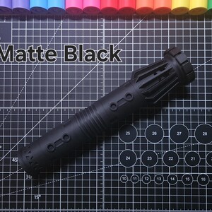 Pu&ograve; includere: Un oggetto cilindrico nero opaco con una superficie strutturata, con dettagli circolari e rettangolari. Il testo "Matte Black" &egrave; visualizzato sopra l'oggetto. Lo sfondo &egrave; un tappetino da taglio nero con linee di griglia e segni di misurazione.