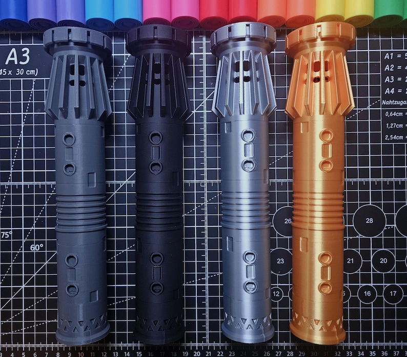 Pu&ograve; includere: Quattro elsi di spada laser stampati in 3D in grigio, nero, argento e oro. Ogni elsa presenta un design dettagliato con sezioni a coste, accenti circolari e una base geometrica. Su un tappetino da taglio nero con misure metriche.