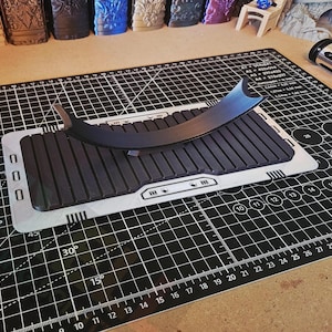 Puede incluir: Un soporte para sable de luz negro y gris con una parte superior curvada, apoyado sobre una esterilla de corte negra y gris. El soporte tiene una base rectangular con l&iacute;neas paralelas. La esterilla de corte tiene un patr&oacute;n de cuadr&iacute;cula y el texto "A3 45 x 30 cm".