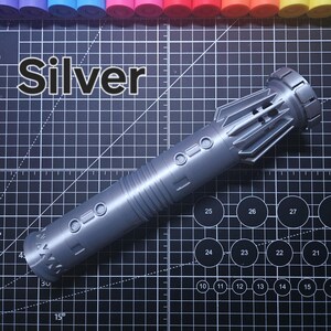Pu&ograve; includere: Un'elica per spada laser argentata con un design cilindrico dettagliato. La parola "Silver" &egrave; stampata a grandi lettere sopra l'elica. L'elica &egrave; su una superficie a griglia nera con cerchi numerati e linee angolate.