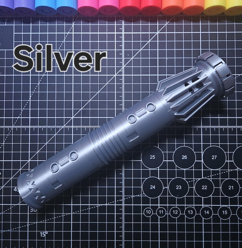 Pu&ograve; includere: Un'elica per spada laser argentata con un design cilindrico dettagliato. La parola "Silver" &egrave; stampata a grandi lettere sopra l'elica. L'elica &egrave; su una superficie a griglia nera con cerchi numerati e linee angolate.